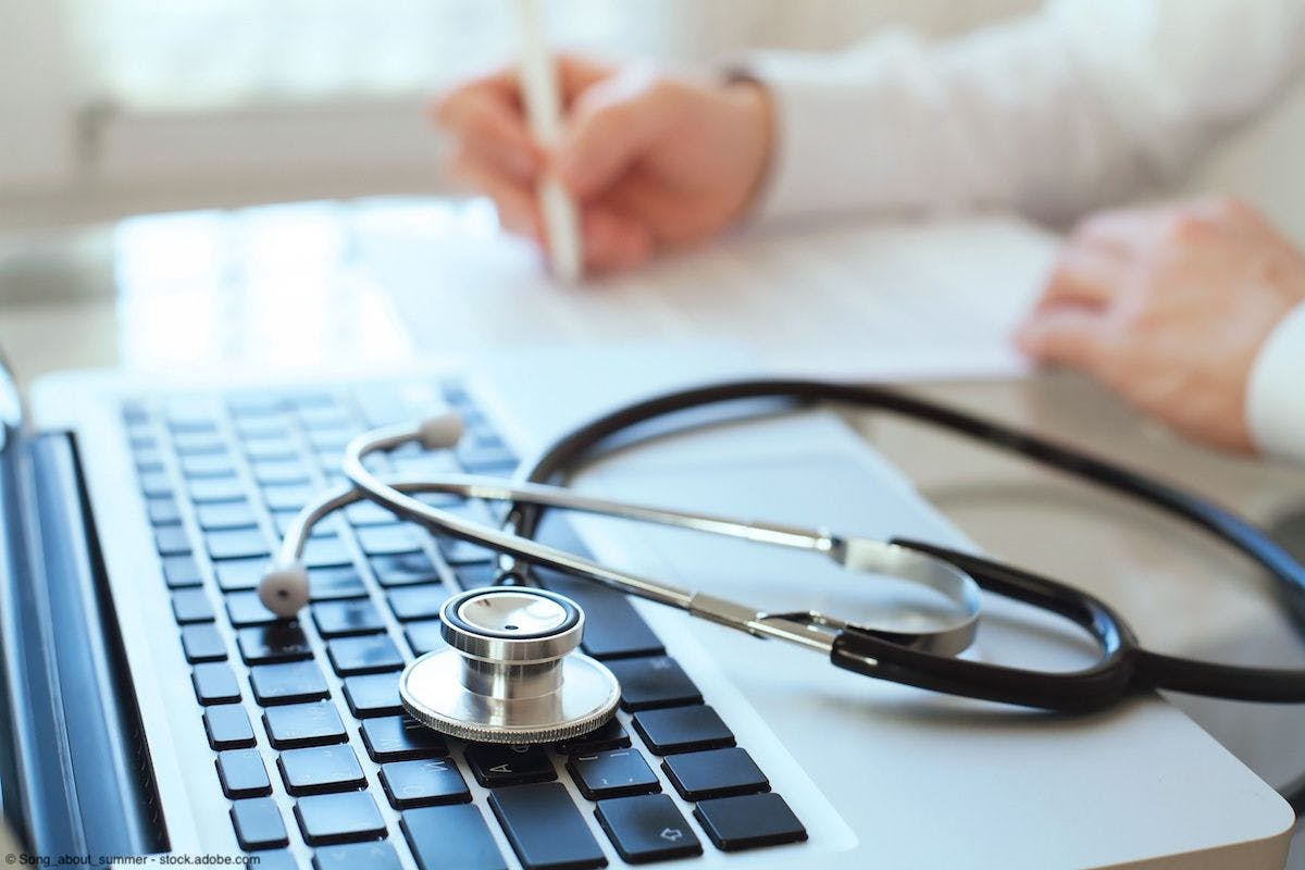 laptop and stethoscope in foreground, doctor's hands writing in blurred background